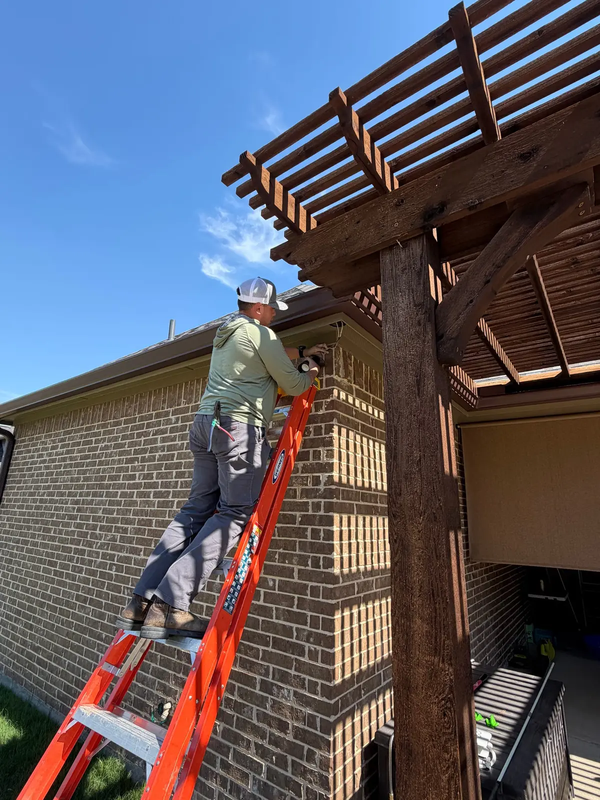 Electrician on an orange extension ladder wiring a new exterior light fixture under the eave beside a cedar pergola at a Midlothian, Texas home